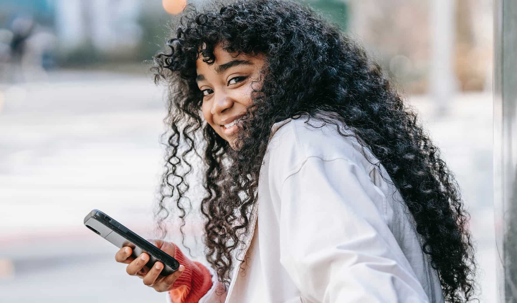 Lachende vrouw met lang krullend haar houdt smartphone vast op straat